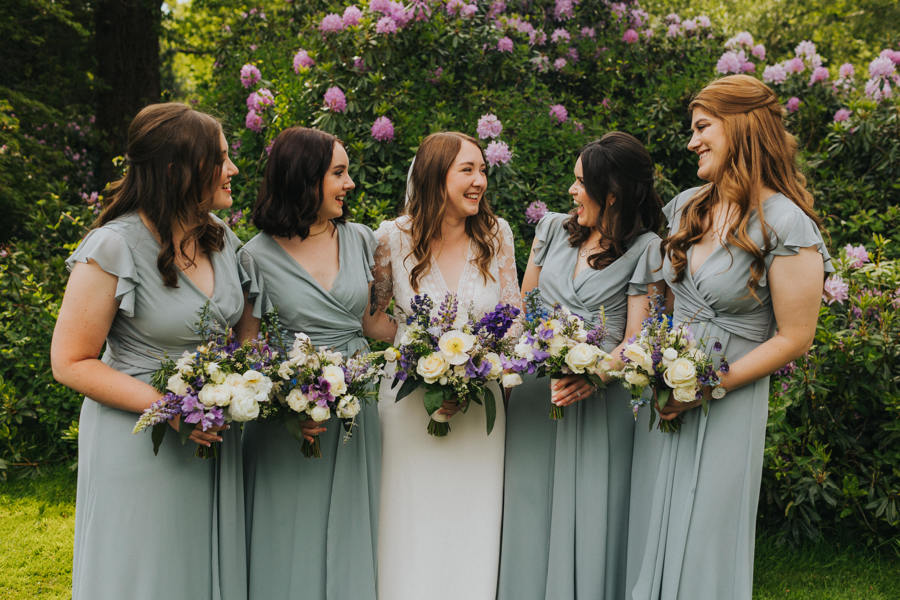 A bride stands in the middle of her four bridesmaids as they hold their bouquets in front of them. They are smiling and laughing at each other and the bridesmaids wear floor length dresses in a green sage tone
