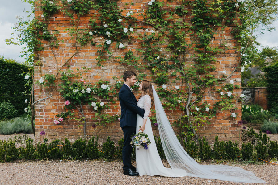 A bride and groom embrace each other in front of a Georgian redbrick Dovecot that has pink climbing roses scrambling up the building