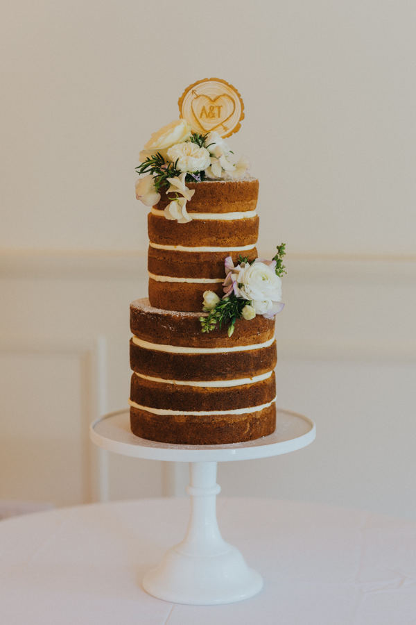 A naked two tier deep wedding cake is positioned on a plain white cake stand with florals and a wooden log topper