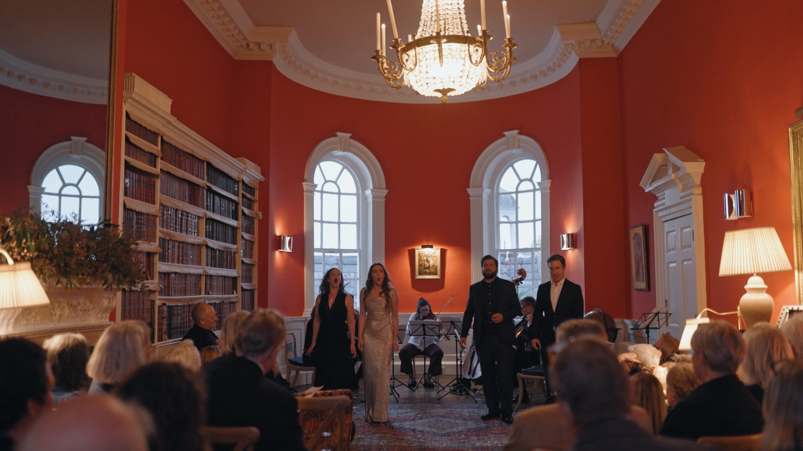 Two men wearing black suits and white shirt stands in a classical Library with two women wearing evening gowns with a quartet of string players playing behind them as they sing opera.