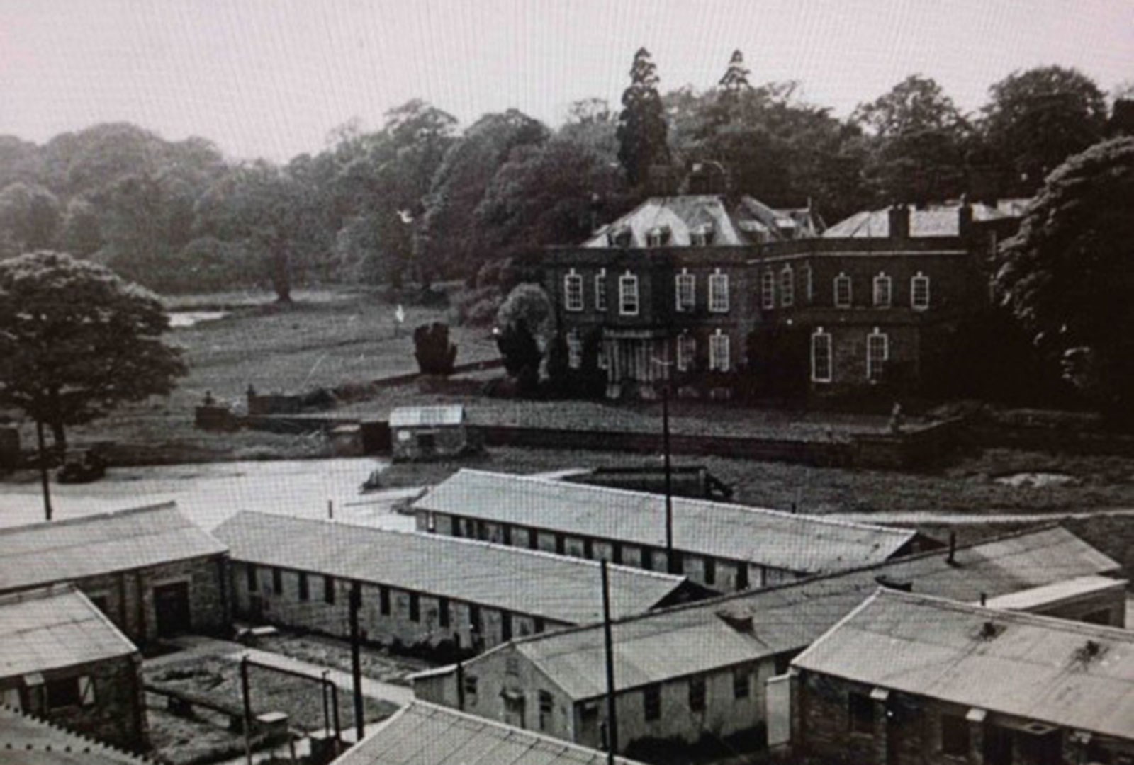 A view of temporary army barracks in the foreground of landscaped gardens with a grand country house in the background