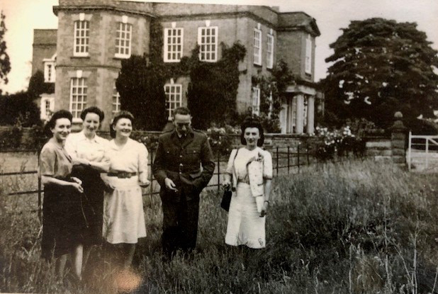 A view of four individuals during the Second World War standing in some gardens with a grand country house in the background