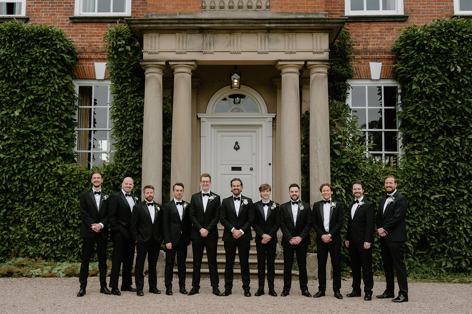 A line up of eleven groomsmen wearing black tie standing outside a Georgian mansion with a Portland Stone portico with their hands clasped in front of them.