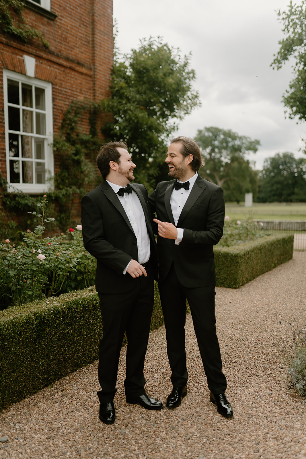 A bridegroom laughs with his best man as they wear black tie standing outside in the gardens of a Manor House