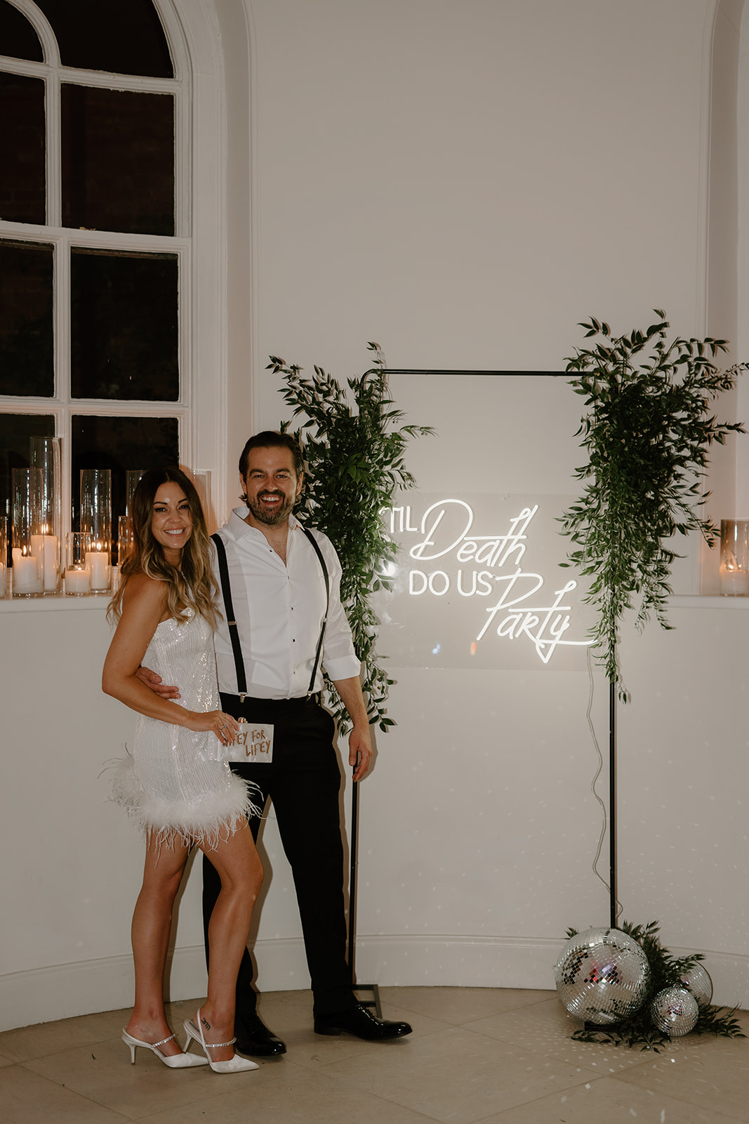 A bride and groom stand proudly next to a neon sign that says 'til death do us party'