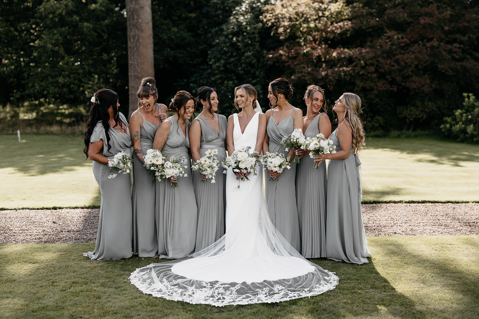 A bride stands in the middle of her bridesmaids wearing sage green dresses and holding white floral bouquets