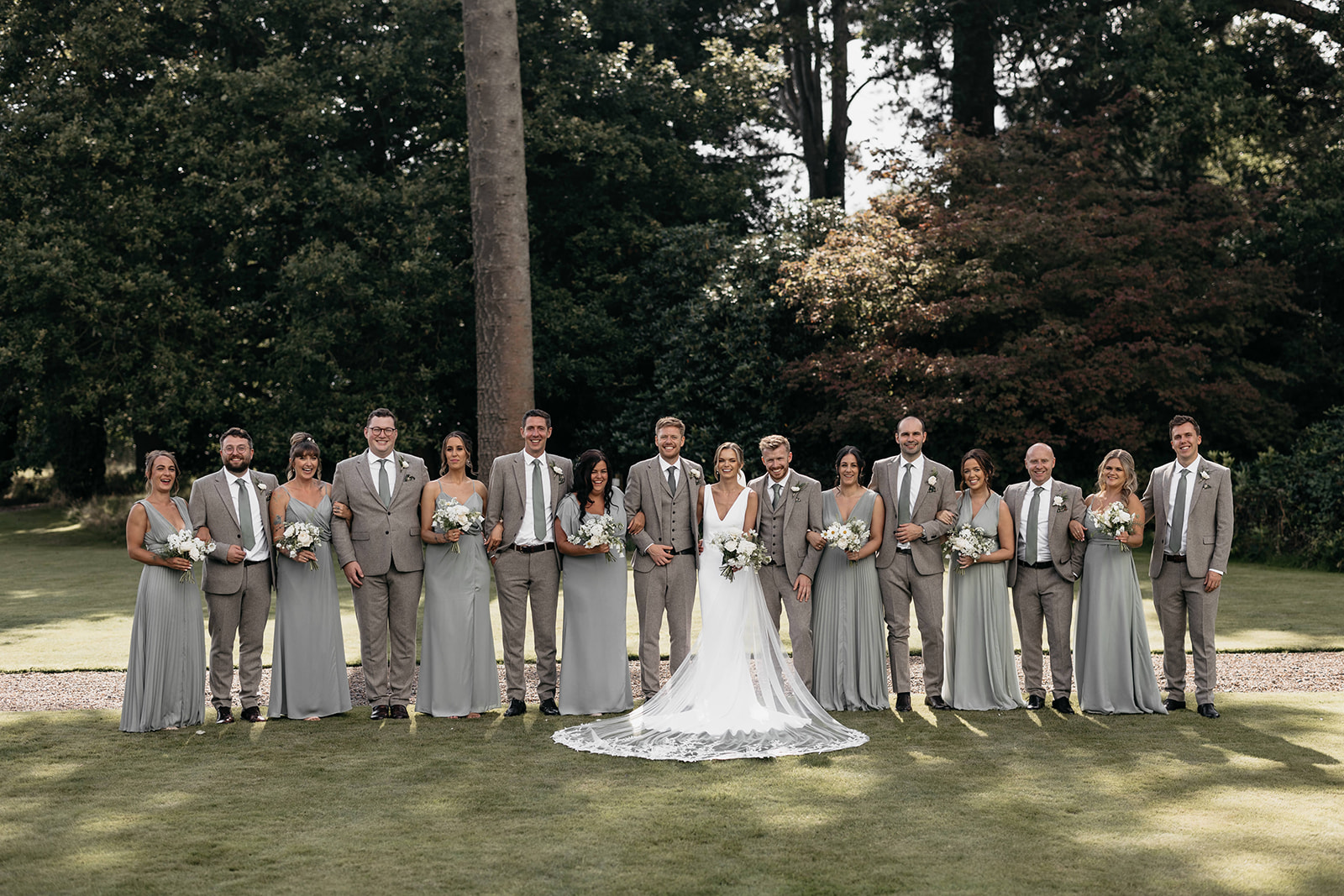 A bride and groom stand in the middle of their bridal party with ushers wearing grey suits and sage green ties and the bridesmaids in green dresses