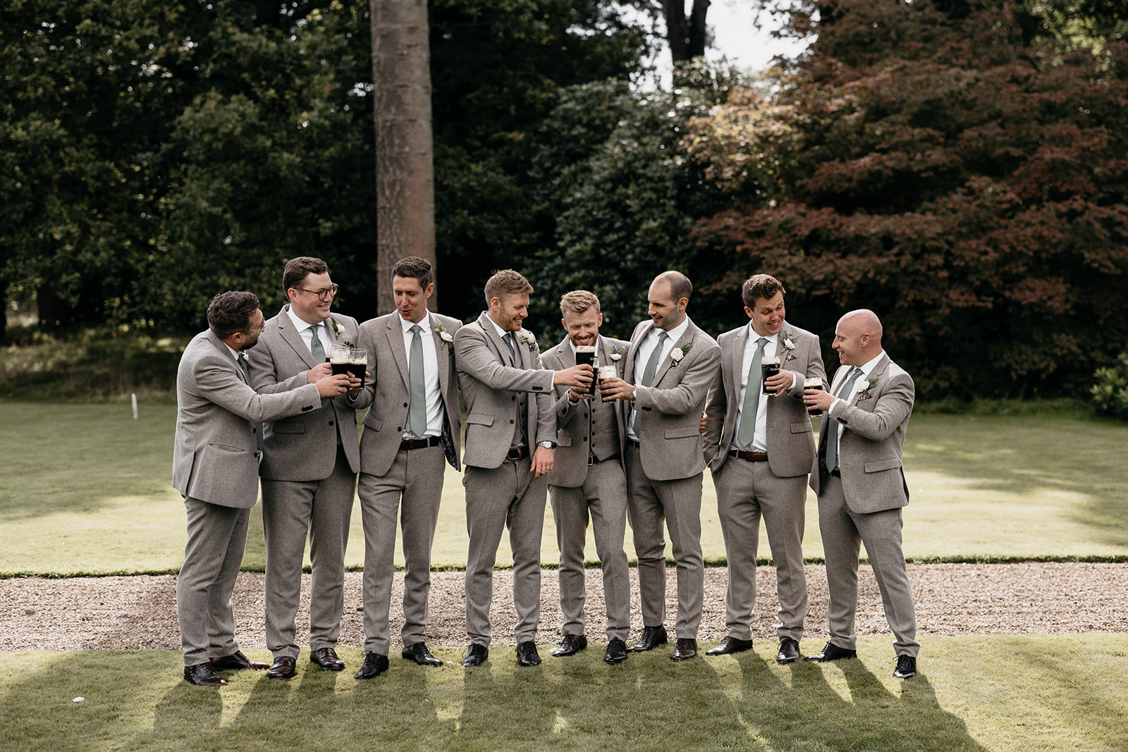 A groom stands in the middle of all of his ushers clinking pints of Guinness whilst wearing grey suits and sage green ties