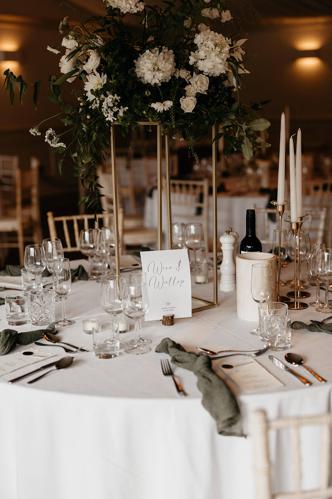 A white and green themed wedding reception table that has sage green linen napkins, cut glassware and elevated floral centrepieces with white candles