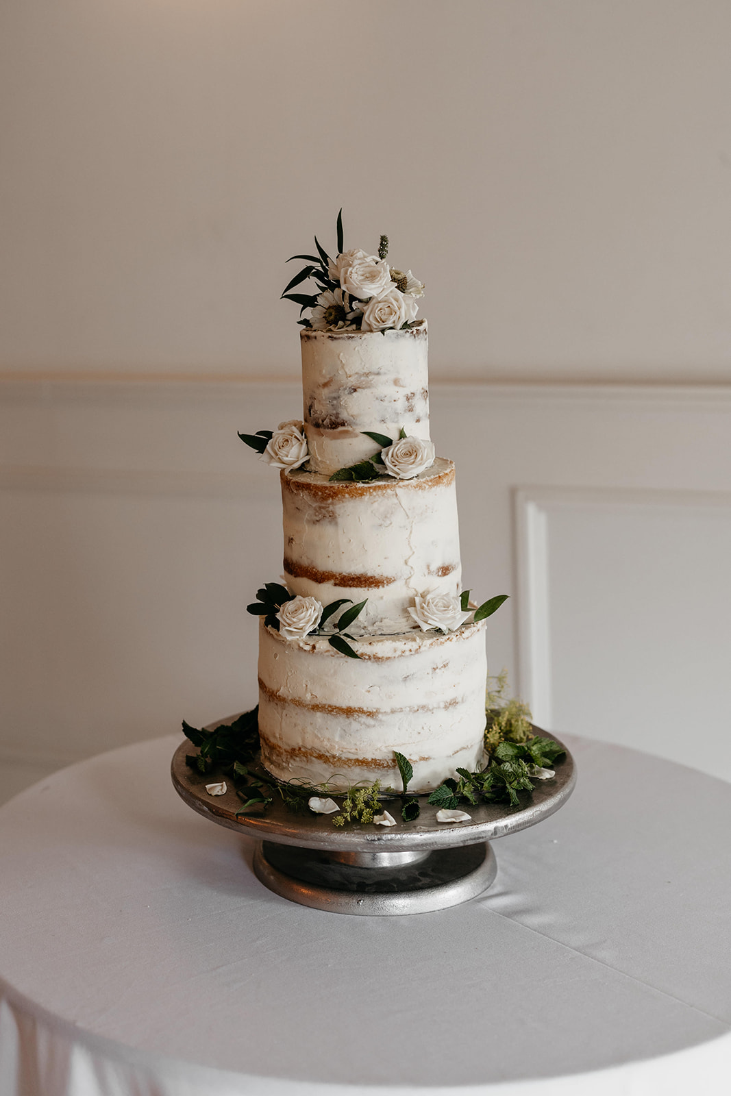 A three tiered naked wedding cake stands on a table covered in a white tablecloth and is garnished in green foliage and white roses