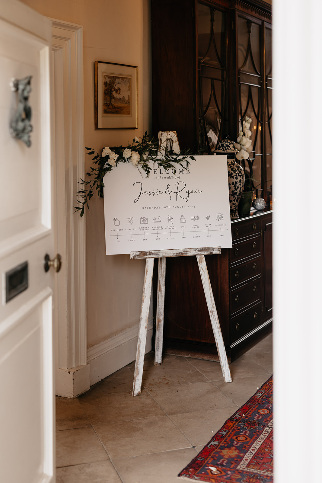 A wedding sign stands propped on an easel in a hallway that has a flagstone floor and red rugs on the floor