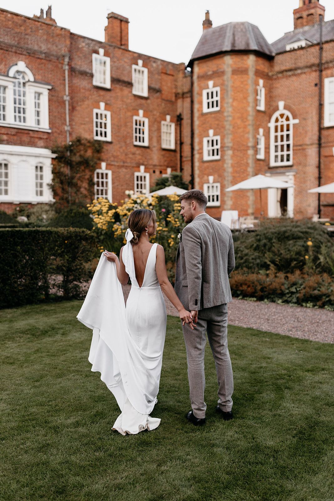 A bride and groom stand outside in a country garden in the middle of summer with a redbrick mansion in the background